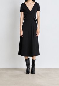 Kobieta ubrana w czarną midi sukienkę z dekoltem w serek, z krótkimi rękawami i zapięciem w talii, w połączeniu z czarnymi skórzanymi botkami sięgającymi do połowy łydki, stojąca na jasnej podłodze.