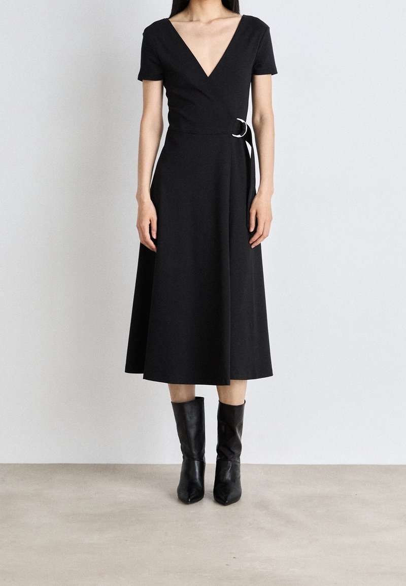 Kobieta ubrana w czarną midi sukienkę z dekoltem w serek, z krótkimi rękawami i zapięciem w talii, w połączeniu z czarnymi skórzanymi botkami sięgającymi do połowy łydki, stojąca na jasnej podłodze.