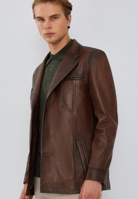 Chaqueta de cuero marrón con una forma estructurada, solapas pronunciadas y un bolsillo con cremallera. Presenta una textura suave y un diseño minimalista.