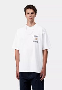 Biała bawełniana koszulka o luźnym kroju, z napisem i logo "Carhartt University" w czerni i pomarańczy na lewej piersi. Srebrny łańcuszek na szyi.