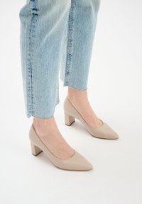 Beżowe skórzane czółenka z ostrym noskiem i słupkowym obcasem. Noszone z jasnoniebieskimi dżinsami z postrzepionymi nogawkami, co podkreśla swobodny styl.