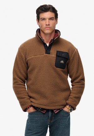Bruine fleece pullover met een zwarte zak, knopenkraag, textuurstof en een geruit overhemd eronder. Bevat een logo op de zak.