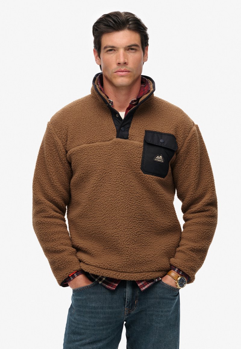 Brown fleece pullover with a black pocket, button collar, textured fabric, and a plaid shirt underneath. Features a logo on the pocket.