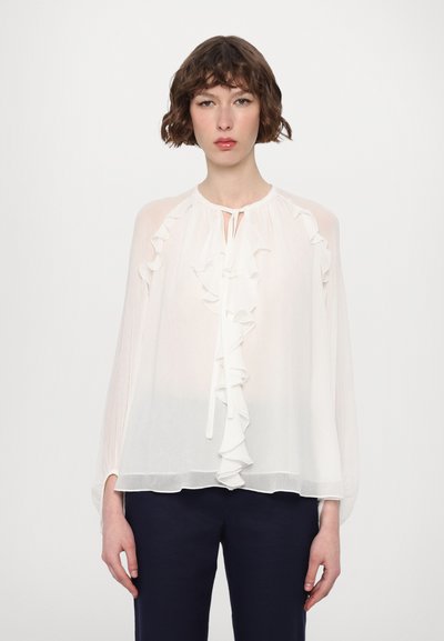 Blouse blanche à manches longues en tissu transparent, présentant un devant froncé et un col rond avec un lien. Associée à un pantalon bleu foncé.