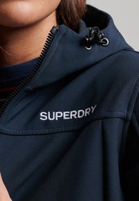 Mörkblå jacka med dragkedja, justerbar huva och vitt "SUPERDRY"-logotyp på kragen. Slät tyg med en mjuk textur.