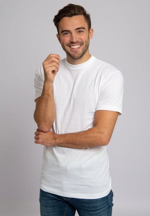 Witte katoenen t-shirt met korte mouwen, ronde halslijn en een gladde textuur, gedragen met blauwe jeans. Het model staat met de armen gekruist.