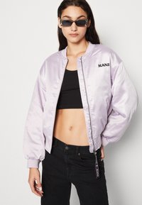 Giacca bomber in satin lilla con logo ricamato nero, design aderente, polsini a coste e chiusura con zip, abbinata a un crop top nero e jeans.