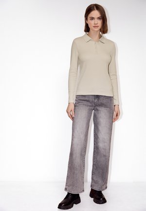 Kobieta ubrana w beżową koszulę z długim rękawem i kołnierzykiem, wyblakłe szare jeansy o prostych nogawkach oraz czarne masywne buty, stojąca na białym tle.