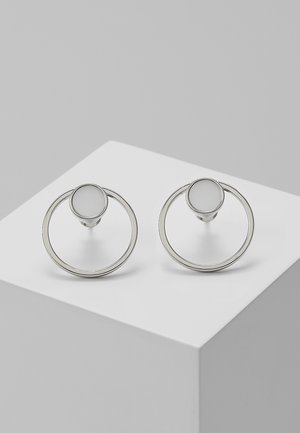 Paire de boucles d'oreilles clous circulaires en argent avec un petit cercle solide fixé au-dessus de plus grandes anneaux ouverts, présentées sur une surface blanche.