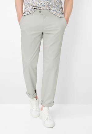 Man met lichtgrijze omgeslagen broek, een bloemenpatroon overhemd in de broek gestopt en witte sneakers, met handen in de zakken.