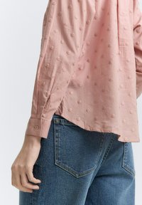 Camicia rosa a trama con motivo a punti in rilievo, maniche lunghe con polsini bottonati, abbinata a jeans in denim blu.