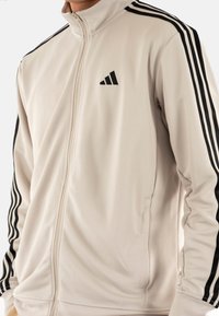 Chaqueta con cremallera en beige claro con franjas laterales negras, fabricada en tela suave, con un pequeño logo negro en el pecho y bolsillos ocultos.