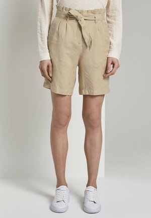 Persona con pantalones cortos beige hasta la rodilla con cinturón anudado, camiseta blanca de manga larga y zapatillas blancas, de pie frente a un fondo liso.