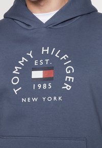 Námořnický modrý mikina s kapucí s předním logem s nápisem "TOMMY HILFIGER," obdélníkovým emblémem v červené, bílé a modré barvě a textem níže.