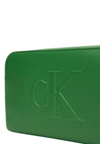 Borsa in pelle verde con logo "CK" in rilievo. Texture liscia, forma rettangolare e dettaglio di cucitura a contrasto lungo i bordi.