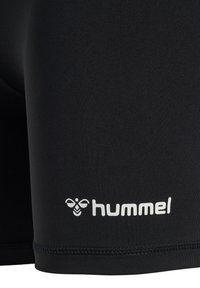 Schwarzer Stoff mit weißem Hummel-Logo und Bienen-Icon, das nahe dem eingenähten Saum von Sportshorts oder Leggings gedruckt ist.