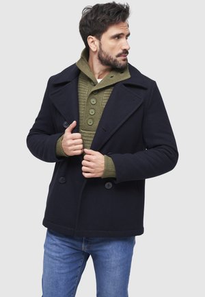 Mies säätää tummaa laivastonsinistä päällystakkia vihreän, napitetun neulepuseron päällä, pukeutuneena sinisiin farkkuihin, katsoen oikealle yksiväristä taustaa vasten.