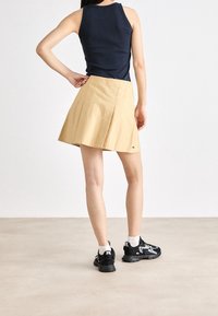 Gonna mini plissettata khaki con cuciture bianco sporco, abbinata a un top senza maniche nero e sneakers nere con dettagli bianchi.