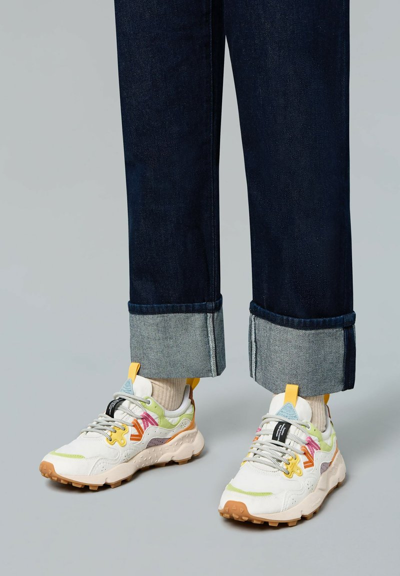 Zapatillas con acentos multicolores, con un upper blanco y gris claro, suela de goma texturizada y cordones visibles. Jeans de mezclilla con los puños enrollados.