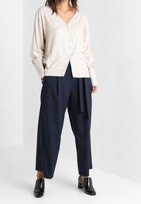 Camicia button-up color beige chiaro con piccoli pois, abbinata a pantaloni ampi blu navy e scarpe nere in vernice. Tessuto morbido e vestibilità comoda.