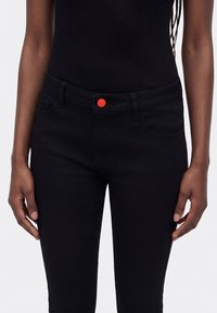 Person iført svarte høytlivsjeans med en knallrød knapp og en svart ribbestrikket ermeløs topp, stående med avslappede armer.
