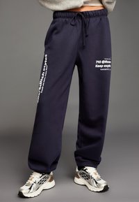 Marinblå mjukisbyxor med dragsko i midjan och vit grafisk text längs vänster ben. Tillverkade av mjukt tyg med en avslappnad passform.