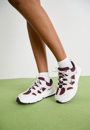 Nogi w białych skarpetkach do kostek oraz w białych i bordowych butach sportowych Nike na zielonej podłodze na tle białej ściany.