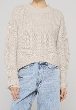 Femme portant un pull beige en grosse maille avec des manches bouffantes, associé à un jean taille haute bleu clair sur un fond uni.