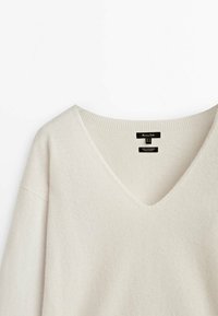 Cremefarbener V-Ausschnitt-Pullover aus Kaschmir mit geripptem Halsbündchen und schwarzen Markenetiketten, die innerhalb des Kragens eingenäht sind.