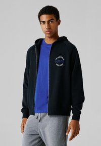 Μαύρο φούτερ με φερμουάρ και κουκούλα με κορδόνι, με ριμπ μανσέτες, φορεμένο πάνω από μπλε μπλουζάκι. Διαθέτει λογότυπο σε άσπρο και μπλε στο στήθος.