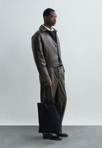 Καφέ δερμάτινο μπουφάν με γιακά, καφέ παντελόνι που ταιριάζει και μαύρη υφασμάτινη τσάντα. Λείος υφασμάτινος χαρακτήρας, μινιμαλιστικός σχεδιασμός και πλαϊνές τσέπες.