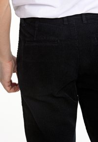 Pantalones de pana negros con canalones verticales, que cuentan con bolsillos traseros y una cintura ajustada. Mano descansando en el costado, exhibidos frente a un fondo neutro.