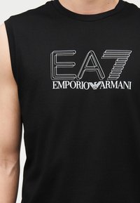 Μαύρο πουκάμισο χωρίς μανίκια με λογότυπο "EA7 Emporio Armani" σε λευκό περίγραμμα και συμπαγές κείμενο στο στήθος, που φοριέται από ένα άτομο.