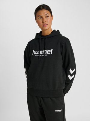 Жена, носеща черен суитшърт и панталони hummel с бяло лого, гледа настрани на светъл едноцветен фон.