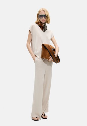 Blondynka w dużych okularach przeciwsłonecznych, beżowym topie bez rękawów, szerokich spodniach, zadrukowanej apasce na szyi, trzymająca brązową zamszową torbę, stojąca z rękami w kieszeniach.