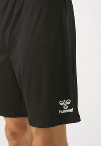 Fekete sport sortot viselő személy, fehér hummel logóval a bal láb szegélye közelében, egyszínű háttér előtt.