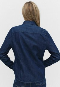 Mörk denimskjorta med lång ärm, knäppkrage och detalj bak på ryggen. Har en skräddarsydd passform och något rundad fåll.