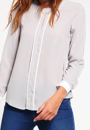 Vrouw draagt een lichtgrijs lang mouwen blouse met witte rand en manchetten, gecombineerd met donkerblauwe spijkerbroek, met één hand bij haar taille.
