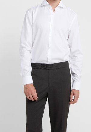 Mężczyzna w białej koszuli wpuszczonej w ciemnoszare spodnie z elastycznym pasem, stojący na jednolitym jasnym tle.