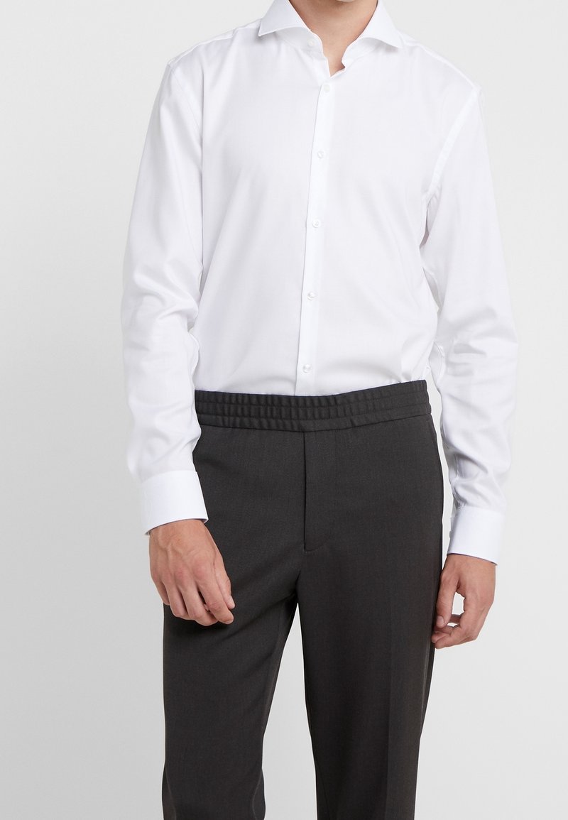Homme portant une chemise blanche rentrée dans un pantalon gris foncé à taille élastique, debout devant un fond clair uni.