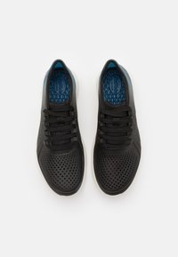 Černé sportovní boty s perforovaným designem, s modrou stélkou a černými tkaničkami, doplněné bílou podrážkou.