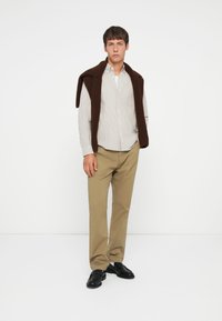 Brun strikket vest over en lysstripet skjorte med knapper, paret med beige bukser og svarte pensko, stående mot en hvit bakgrunn.