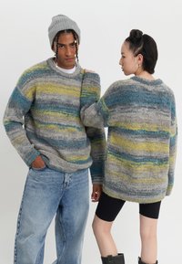 Teksturalni, oversize sivi pulover s plavim i žutim prugama; uparen s trapericama. Sivi beanie. Model ima kratku, stiliziranu kosu.