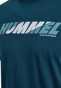 Teal bomullströja med en djärv vit och ljus teal grafisk text, "HUMMEL," där "ACTIVE SPORTSWEAR" står i mindre teckenstorlek nedanför.