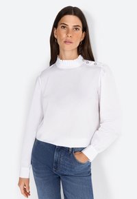 Blouse blanche à manches longues avec un col volant haut, des détails de boutons sur l'épaule et des poignets ajustés. Tissu lisse et léger.