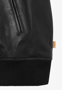 Zwarte leren jas met een geribbelde zwarte brei tailleband en ritszakdetails, met een klein oranje merklabel aan de zijkant.