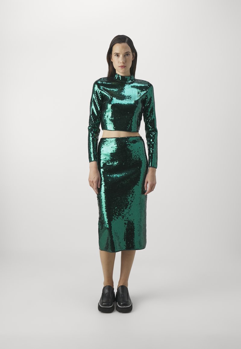 Kobieta stoi przodem, ubrana w błyszczący zielony top z cekinami i długimi rękawami, do którego dobiera midi spódnicę w tym samym kolorze oraz czarne buty.