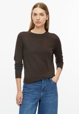 Vrouw met steil lichtbruin haar, gekleed in een donkerbruine trui met lange mouwen en een blauwe spijkerbroek, staand met één hand in haar zak.