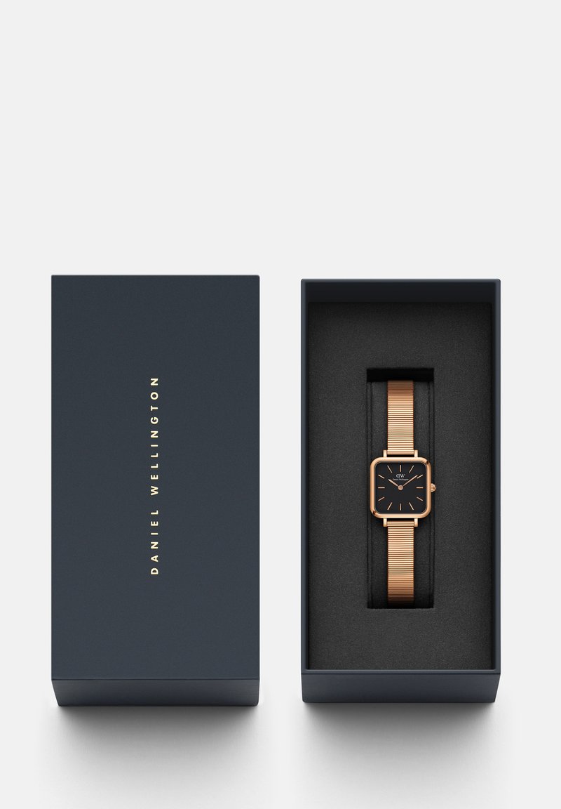 Złoty prostokątny zegarek z czarną tarczą i siateczkowym paskiem w granatowym pudełku wyłożonym czarną pianką. Prosty, nowoczesny design, bez dodatkowych zdobień.