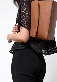 Personne portant un haut en dentelle noire et un pantalon, porte un sac à dos structuré en cuir marron sur une épaule.